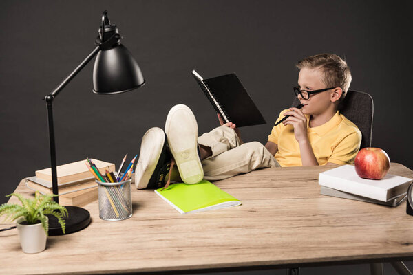schoolboy in eyeglasses doing homework with legs on table with books, plant, lamp, colour pencils, apple, clock and textbook on grey background 
