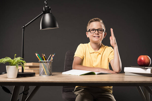 schoolboy in eyeglasses sitting with raised hand at table with books, plant, lamp, colour pencils, apple and textbook on grey background 