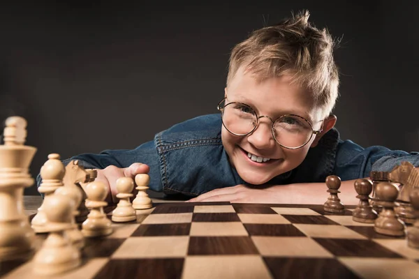 smiling little boy in eyeglasses looking at camera while sitting at table with chess board isolated on grey background