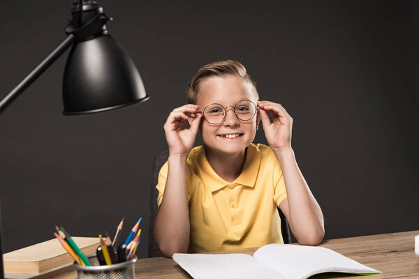 smiling schoolboy adjusting eyeglasses and sitting at table with lamp, colour pencils, books and textbook on grey background 