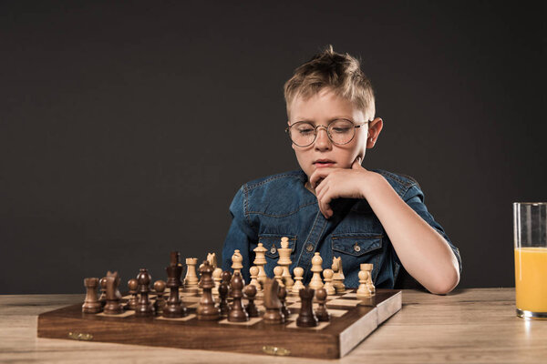 pensive little boy sitting at table with chess board and glass of juice on grey background 