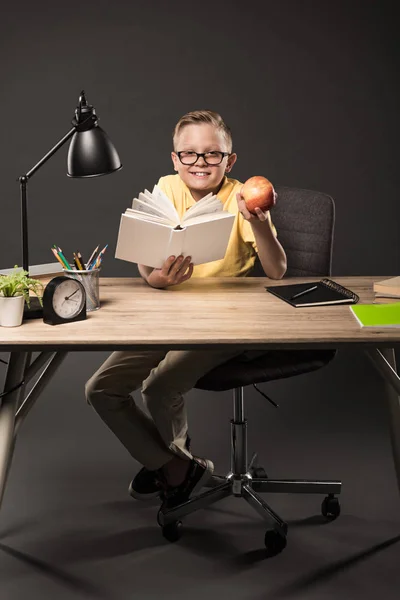 Colegial Sonriente Anteojos Sosteniendo Manzana Libro Lectura Mesa Con Libros — Foto de Stock