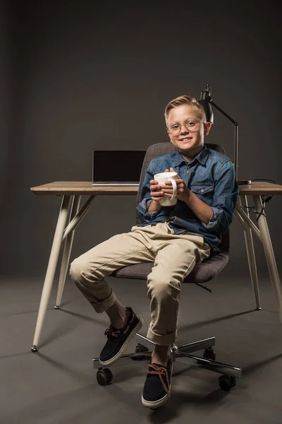 戴眼镜的小男孩拿着咖啡杯 坐在桌子旁边的椅子上 带着笔记本电脑和灯在灰色背景下 — 免费的图库照片