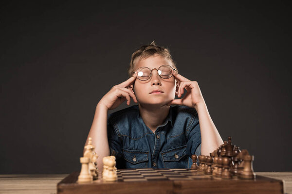 thoughtful little boy with closed eyes in eyeglasses holding fingers on temples at table with chess board on grey background 