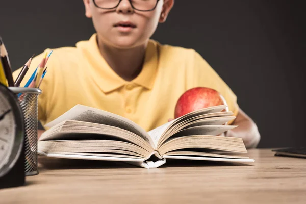 Gambar dipotong anak sekolah dalam kacamata makan apel dan membaca buku di meja dengan pensil warna dan jam di latar belakang abu-abu — Stok Gambar Gambar Dipotong Anak Sekolah Dalam Kacamata Makan Apel Dan Membaca Stok Lukisan
