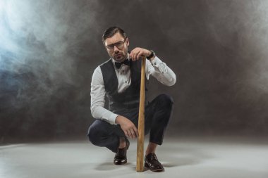 serious stylish man in eyeglasses crouching with baseball bat and looking at camera on black clipart