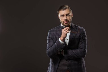 takım elbise ve papyon gözlük tutan ve siyahta izole kamera bakarak yakışıklı adam