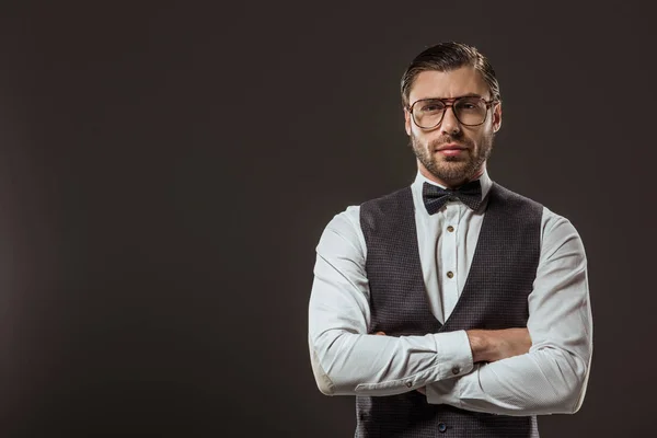 handsome stylish man in bow tie and eyeglasses standing with crossed arms and looking at camera isolated on black