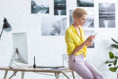 gülümseyen kadın freelancer grafik tablet ve ev ofis bilgisayarı ile tablo yakınındaki smartphone kullanarak yan görünüm 