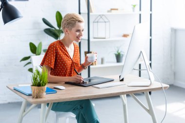 kadın freelancer grafik tablet bilgisayar ile masada çizim kahve fincanı ile gülümseyen 