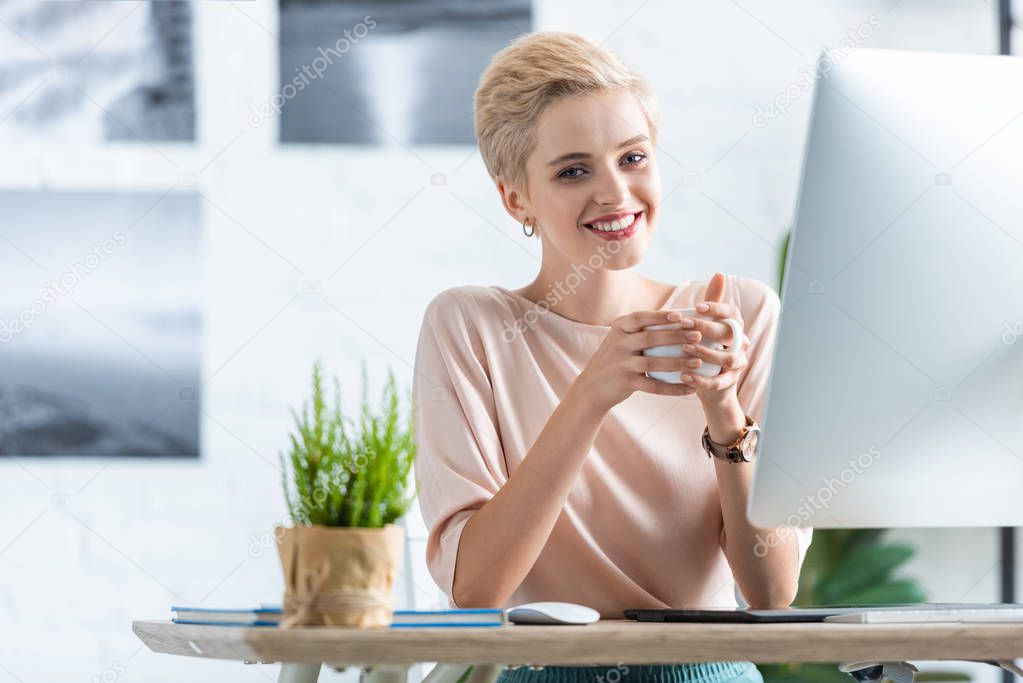 smiling businesswoman holding cup of coffee at table with graphic tablet and computer in office 