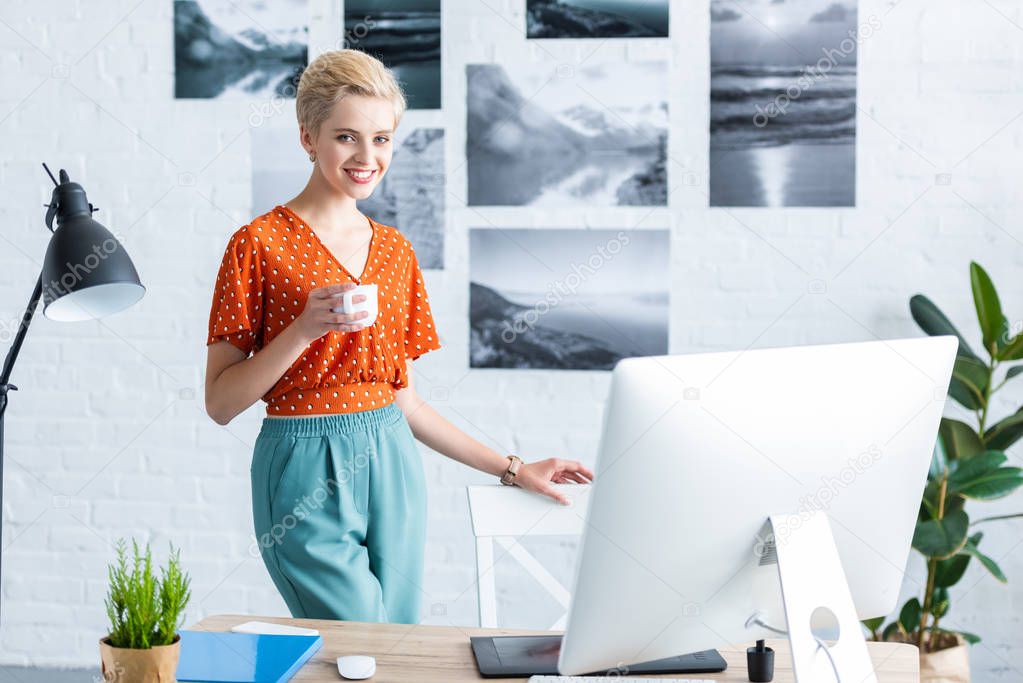 stylish female freelancer holding cup of coffee near table with graphic tablet and computer in home office 