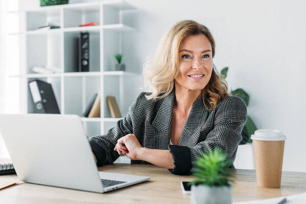 привлекательная деловая женщина сидит за столом с ноутбуком и смотрит в сторону в офисе
