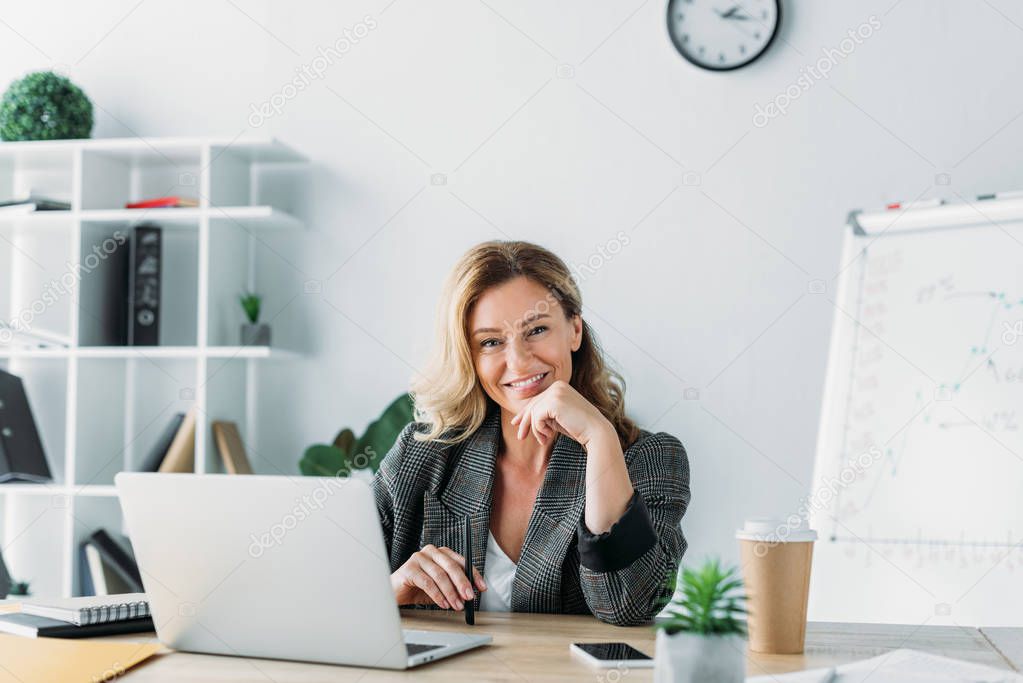 attractive businesswoman sitting at table with laptop and looking at camera in office