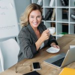 Привлекательная деловая женщина, сидящая с чашкой кофе за столом в офисе Привлекательная деловая женщина, сидящая с чашкой кофе за столом в офисе