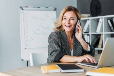 smiling beautiful businesswoman talking by smartphone in office and looking away clipart