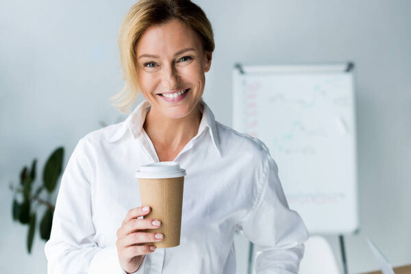 улыбающаяся привлекательная деловая женщина с одноразовой чашкой кофе в офисе и смотрящая в камеру
