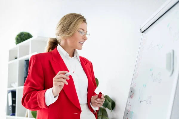 有吸引力的女商人在红色夹克站立在白板附近在办公室和藏品标记 — 图库照片