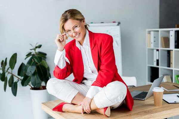 привлекательная деловая женщина сидит на столе и смотрит в камеру в офисе
