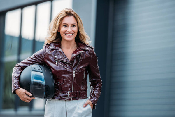 smiling woman in leather jacket holding motorcycle helmet on street and looking away