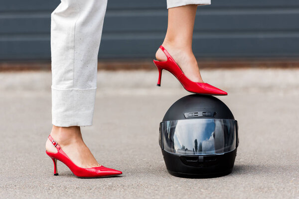 обрезанное изображение женщины на красных каблуках, надевающей ногу на мотоциклетный шлем на улице
 