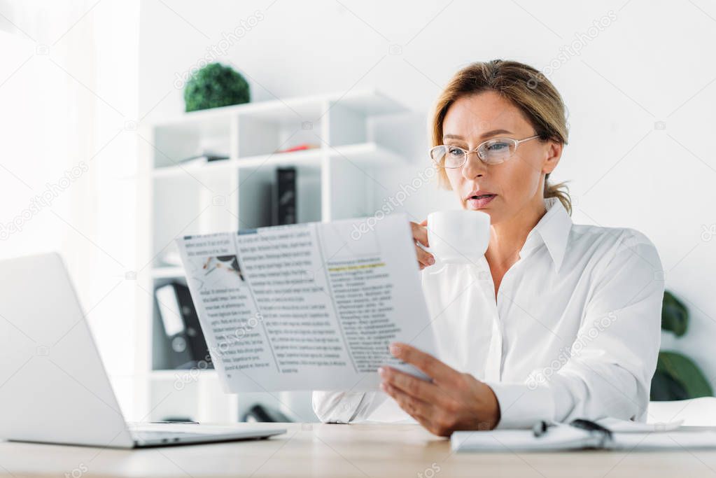 attractive businesswoman drinking coffee and reading newspaper in office