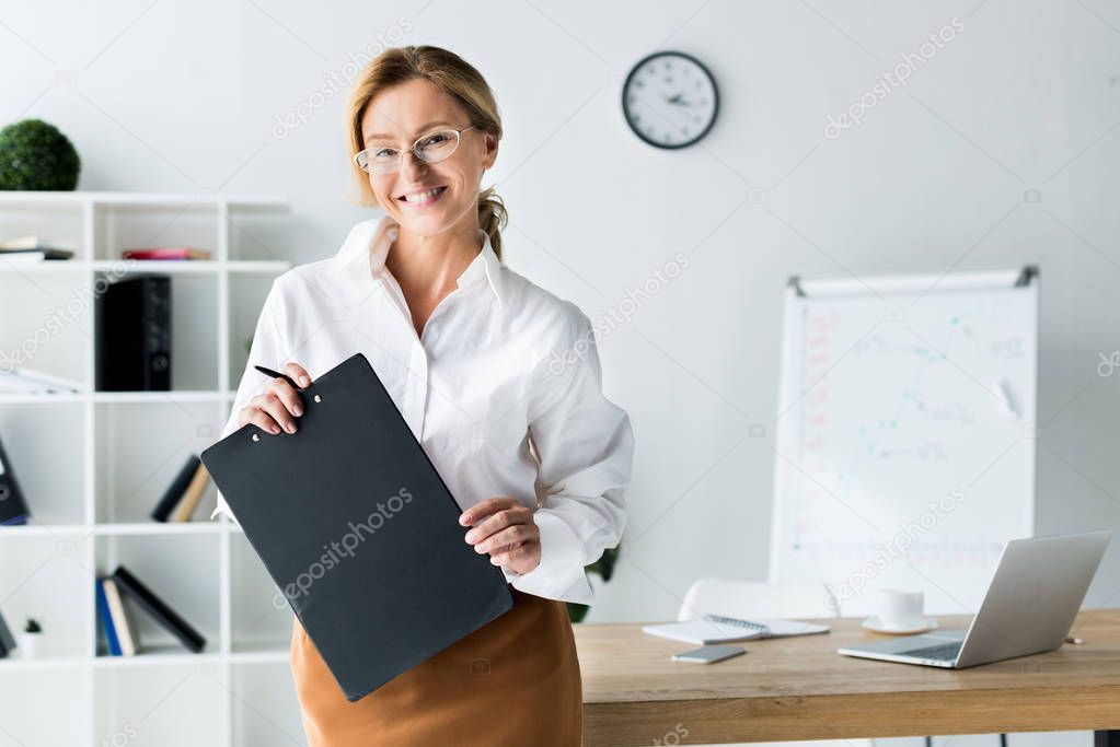 smiling attractive businesswoman holding clipboard and looking at camera in office