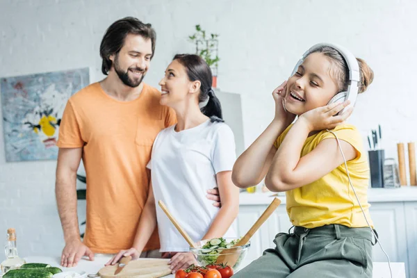 Niña Sonriente Auriculares Sentados Mesa Mientras Sus Padres Abrazan Fondo —  Fotos de Stock