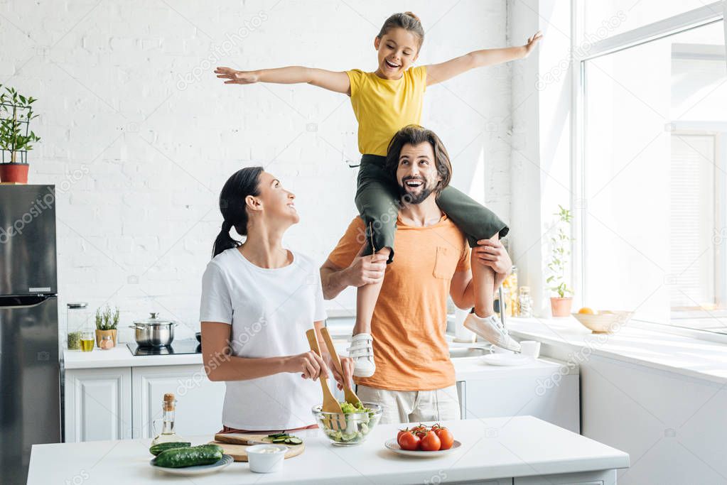 beautiful young woman preparing salad while her daughter riding on shoulders of husband at kitchen