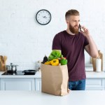 Bonito barbudo jovem falando por smartphone e olhando para longe na cozinha