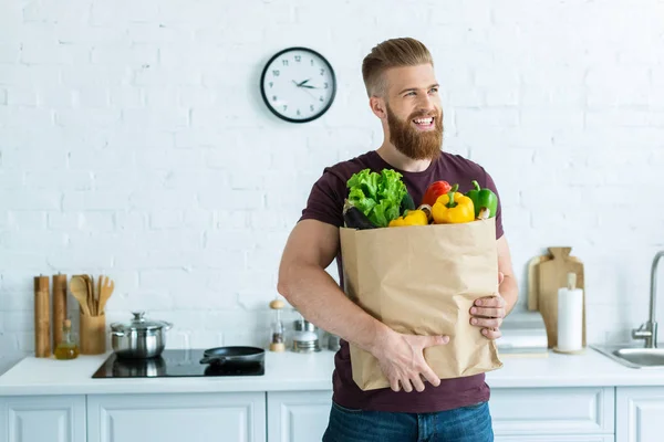 Красивий Усміхнений Молодий Чоловік Тримає Продуктову Сумку Овочами Дивиться Кухню — стокове фото