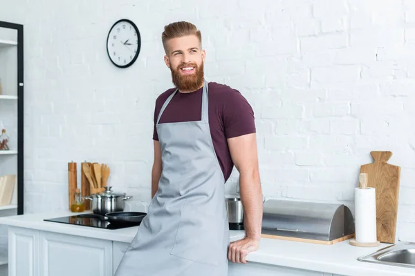 ハンサムなひげを生やした若い男が笑みを浮かべて キッチンに目をそむけるのエプロン — ストック写真