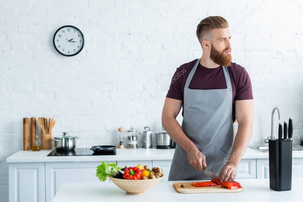 handsome bearded young man in apron looking away while cooking in kitchen