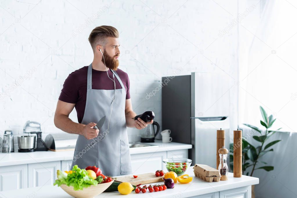 handsome bearded man in apron listening music in earphones and looking away while cooking in kitchen 