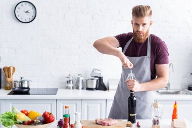 yakışıklı delikanlı şişe şarap açma ve mutfakta yemek yaparken kameraya bakarak önlük sakallı