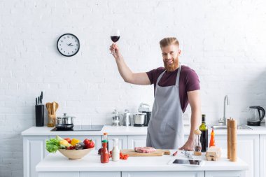 yakışıklı delikanlı kırmızı şarap tutarak ve mutfakta yemek yaparken kameraya gülümseyen önlük sakallı