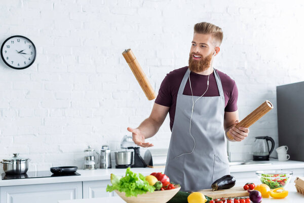 handsome smiling bearded man in apron listening music in earphones while cooking in kitchen