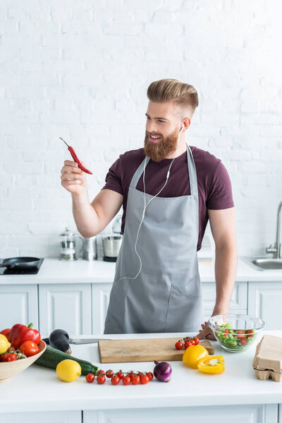 handsome smiling bearded man in apron listening music in earphones and holding chili pepper while cooking in kitchen