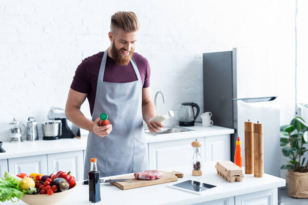 handsome smiling bearded man in apron cooking delicious steak in kitchen