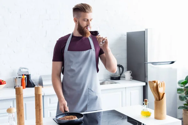 赤ワインを飲みながらフライパンでステーキを調理エプロンでハンサムな若い男  — 無料ストックフォト