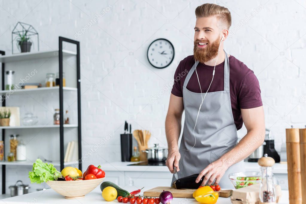 handsome smiling bearded man in apron and earphones cooking vegetable salad in kitchen