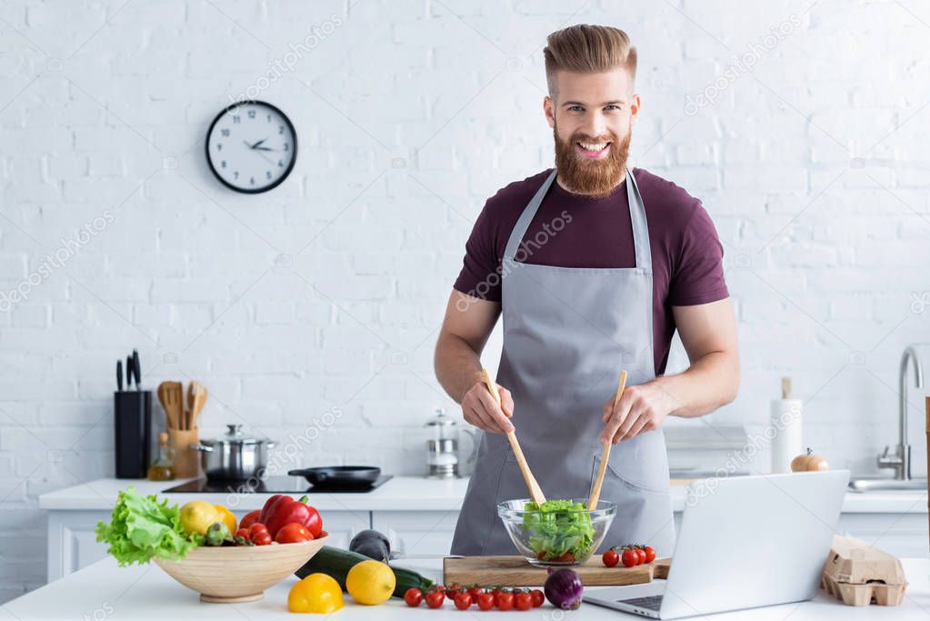 handsome bearded man in apron cooking vegetable salad and smiling at camera