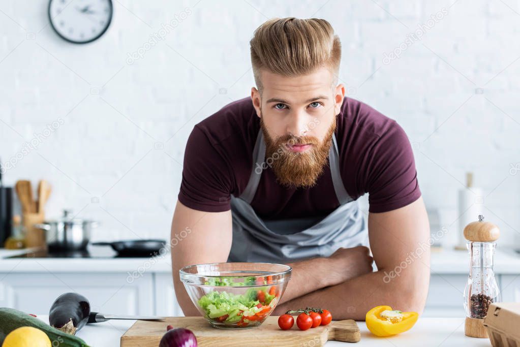 handsome bearded man in apron cooking vegetable salad and looking at camera
