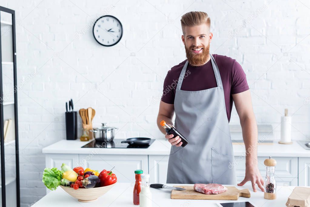 handsome bearded man in apron cooking steak and smiling at camera in kitchen 