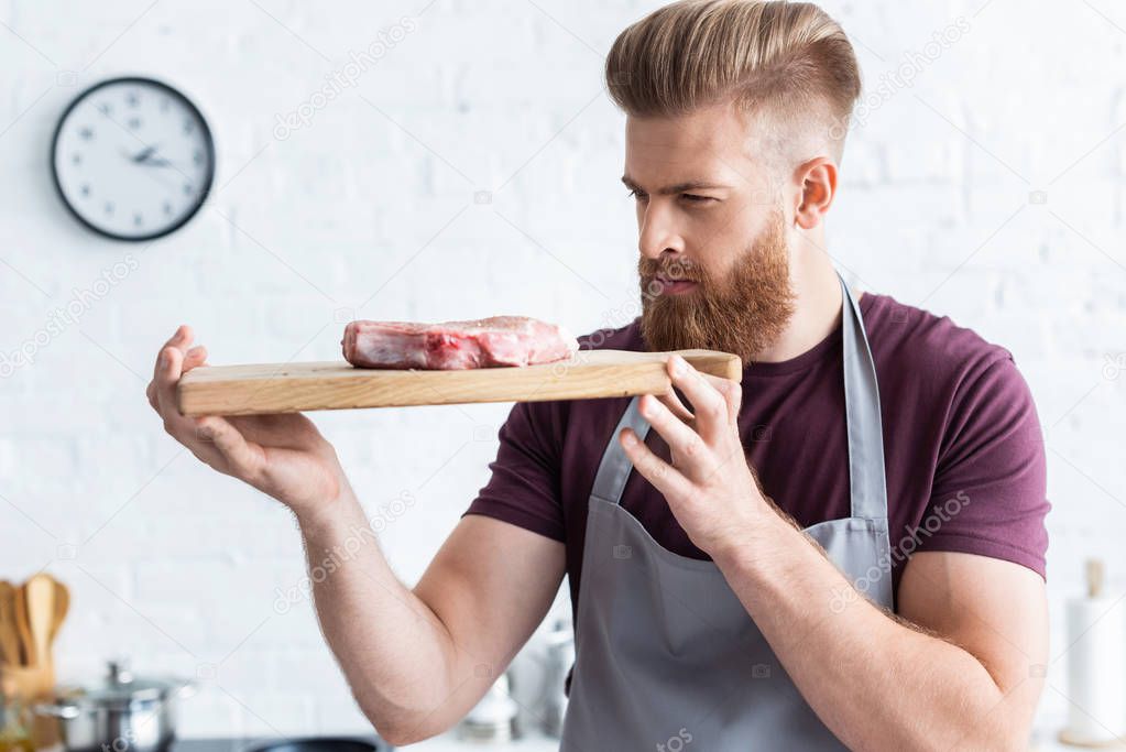 handsome bearded young man holding wooden cutting board with delicious raw beef steak