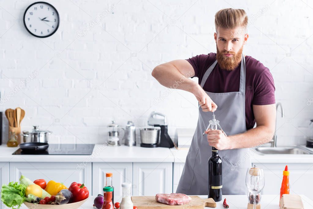 handsome bearded young man in apron opening bottle of wine and looking at camera while cooking in kitchen