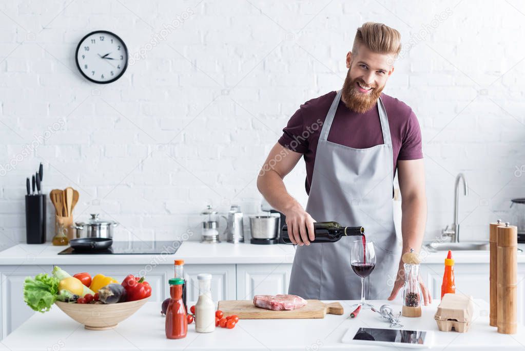handsome bearded young man in apron pouring wine and smiling at camera while cooking in kitchen