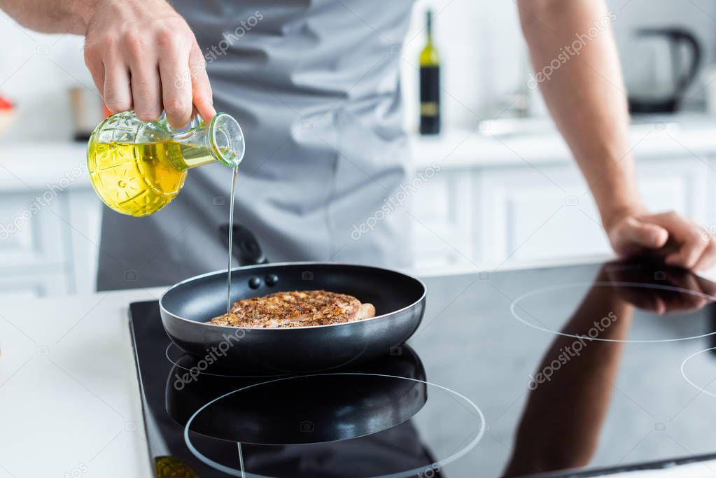 cropped shot of man in apron pouring oil while cooking steak on frying pan