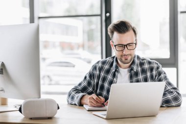 handsome young programmer in eyeglasses taking notes while working with laptop and desktop computer clipart