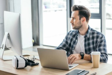 handsome young programmer using laptop and desktop computer at workplace clipart